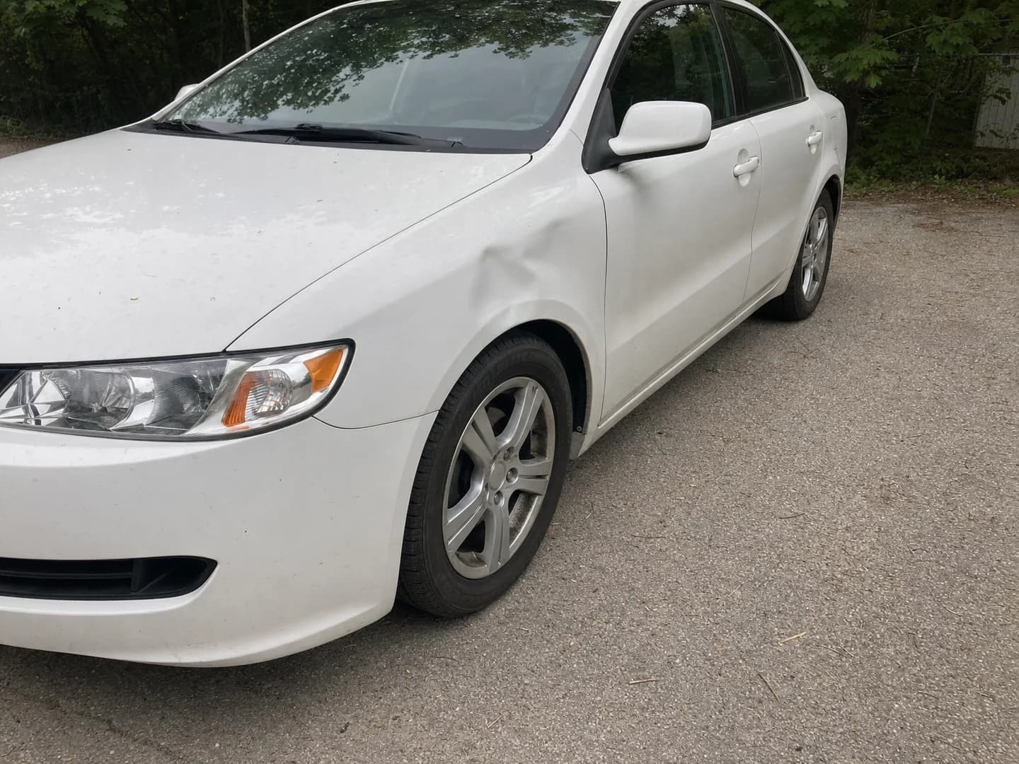 White sedan with a dent in the front fender on a gravel parking lot
